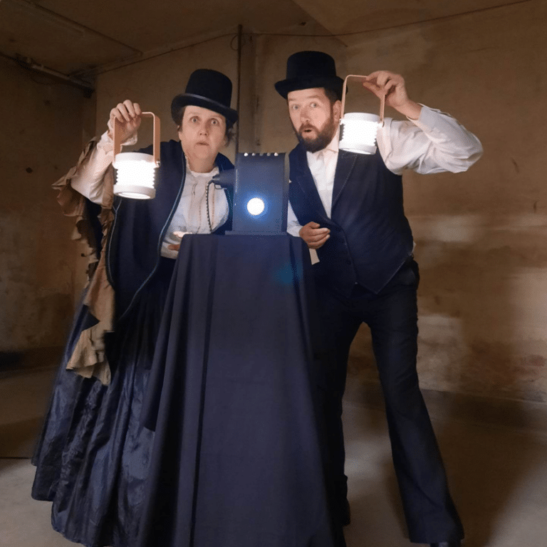 Two performers in Victorian costume holding lanterns beside projector