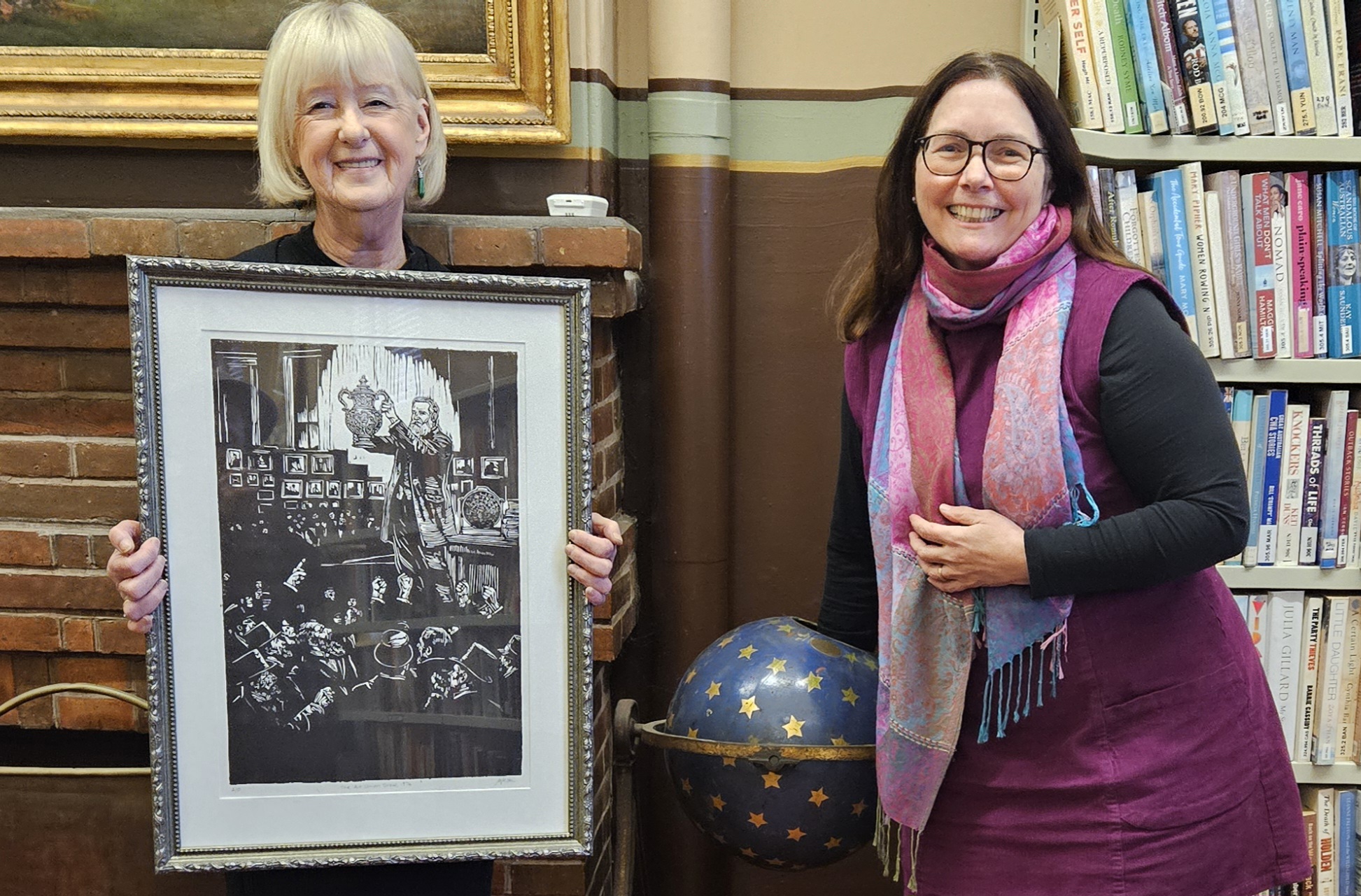 Two women smiling holding framed artwork in a library