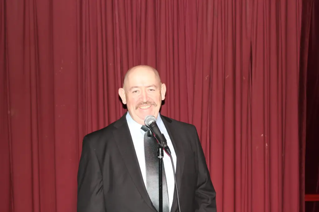 Smiling bald man with moustache speaking at microphone on stage