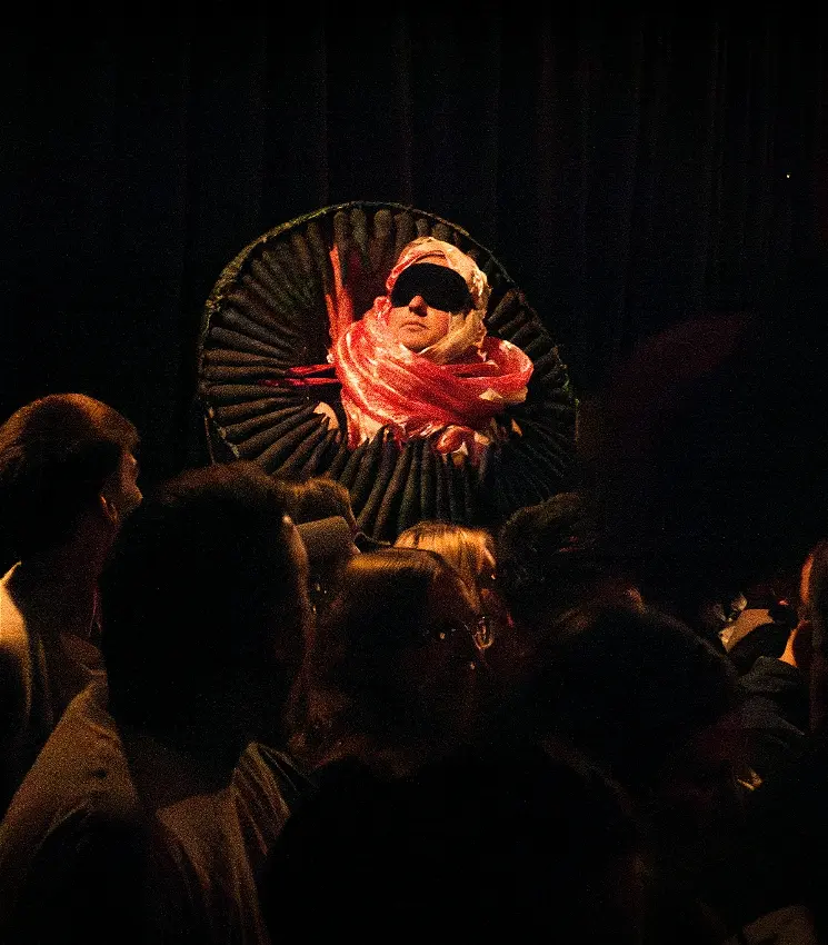 Blindfolded performer in peacock chair above dark crowd