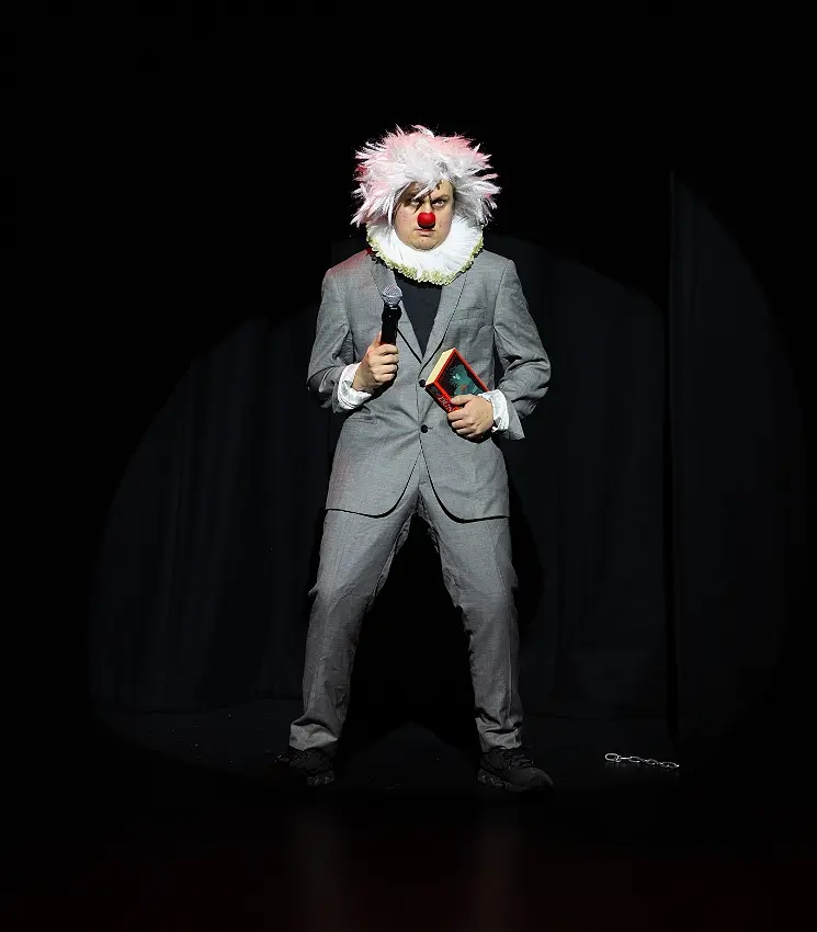 Clown in grey suit holding microphone and book on stage